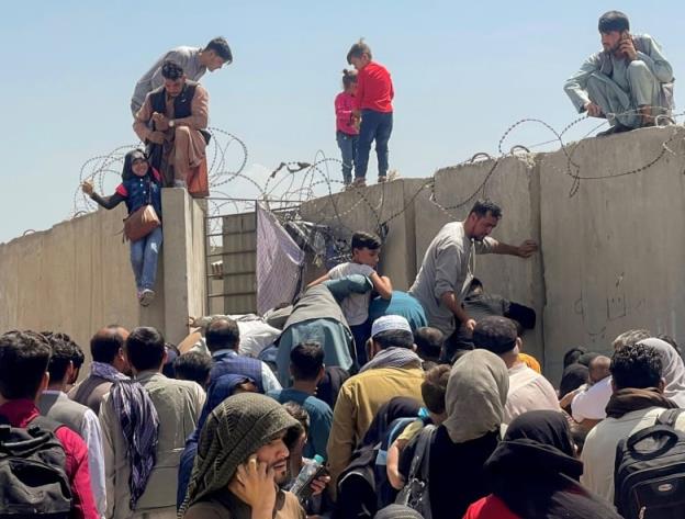 Men, women and children clamber over a co<em></em>ncrete wall topped with razor wire. 