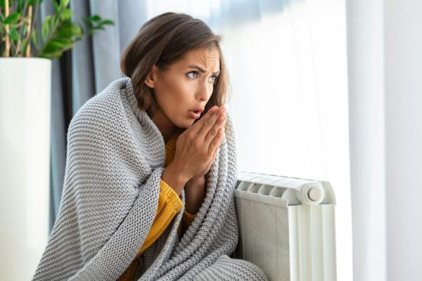 woman in grey blanket huddled over radiator, looks cold