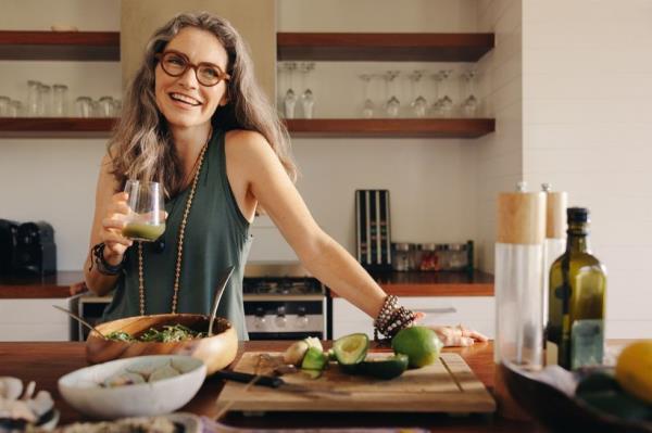Healthy senior woman smiling while holding some green juice and making salad in her kitchen.