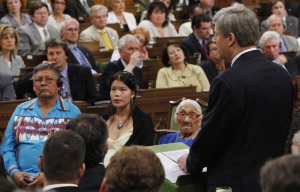 A gallery of seated observers and a man in a dark suit seen from behind, standing at a lectern.