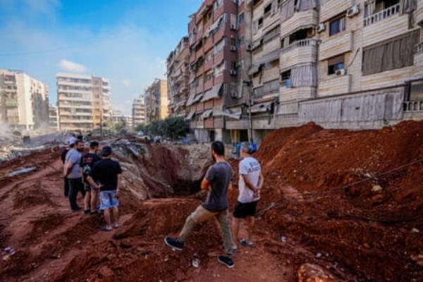 People standing near a tunnel visible in red raised earth beside bomb-damaged buildings