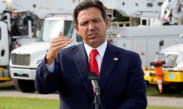 Ron DeSantis gestures during an outdoor news conference. There are parked vehicles in the background