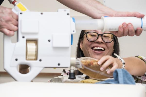 A NASA food scientist smiles as she rehydrates a food pouch using the Mini Potable Water Dispenser during a demo<em></em>nstration at Johnson Space Center. The scientist holds the food pouch steady while another person manually operates the handheld device, which is designed to dispense water for preparing meals and maintaining hygiene on the Gateway lunar space station.