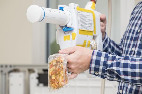 The image shows a close-up of an engineer's hands as they operate a Mini Potable Water Dispenser. The engineer is holding the white, handheld device, which features a large, graduated syringe-like compo<em></em>nent and is marked with various yellow labels and instructions. The engineer is rehydrating a vacuum-sealed food pouch, which he is holding in one hand while the other hand operates the dispenser. The food inside the pouch appears to be a mix of ingredients. The engineer is wearing a blue checkered shirt.