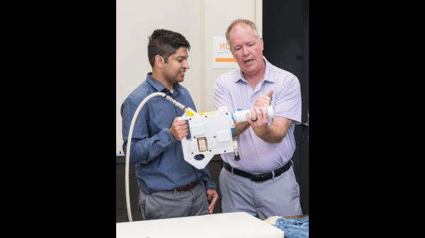 The image shows two individuals standing side by side as they demo<em></em>nstrate the Mini Potable Water Dispenser. The person on the left, dressed in a blue shirt, is holding the device, while the person on the right, wearing a light purple shirt, is explaining how to use it. The person on the right is gesturing with his hands, explaining the device's functio<em></em>nality or the process of rehydrating food. The device is co<em></em>nnected to a hose and features a large, graduated syringe-like component.