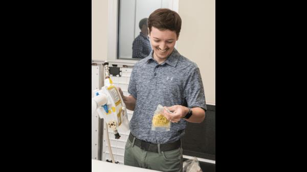The image shows a close-up of a person filming a demo<em></em>nstration with a smartphone. The scene being recorded involves the use of a Mini Potable Water Dispenser to rehydrate a vacuum-sealed food pouch. The person's hand is holding the device while directing water into the pouch, which is positio<em></em>ned on a table. The smartphone's screen is clearly visible in the foreground, displaying a live video of the demo<em></em>nstration in progress