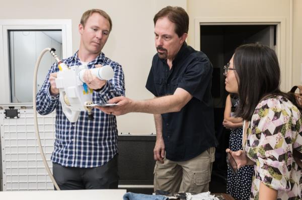 The image shows a group of three people engaged in a demo<em></em>nstration of the Mini Potable Water Dispenser. The person on the left, wearing a checkered shirt, is holding the device and showing its operation to the others. The person in the center is closely examining a vacuum-sealed food pouch being held by the person with the dispenser, while pointing or assisting in the demonstration. The person on the right, wearing glasses and a floral-patterned top, is attentively observing the process, holding a smartphone, possibly ready to capture the demonstration.