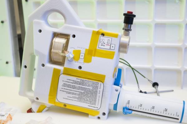 The image shows a close-up of the Mini Potable Water Dispenser prototype. The device is primarily white with yellow tape on it and has several instructio<em></em>nal labels on its surface. It features a prominent graduated syringe-like compo<em></em>nent for precise water measurement, marked in milliliters, and a black push button at the top. The device has a handle on one side for easy portability. The background co<em></em>nsists of a white grid pattern, which helps draw attention to the device. Partially visible in the foreground are vacuum-sealed food pouches, indicating the device's use for rehydrating food.
