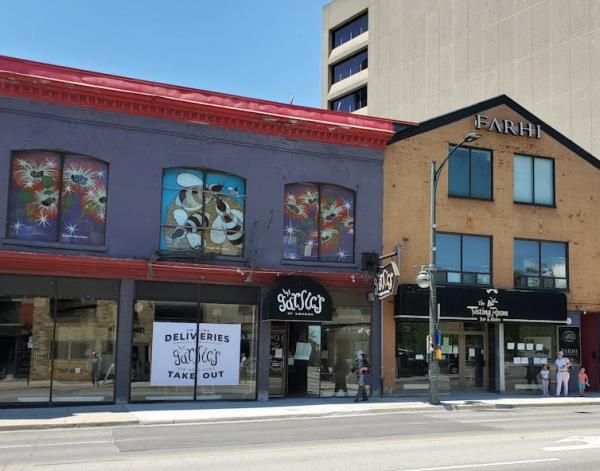 A consistent façade of two-storey buildings on Richmond Street in London, Ontario, Canada housing multiple restaurants. A person walks by wearing a mask, along with a young family not far behind. A large sign hangs in one of the ground floor windows advertising delivery and takeout from Garlic’s restaurant.