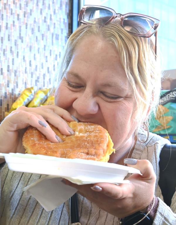 Rosalie Durso biting into a jelly doughnut with bacon, egg, and cheese at Manhattan Best Deli & Grill, New York. Seen with long gray nails and sunglasses on top of her head.