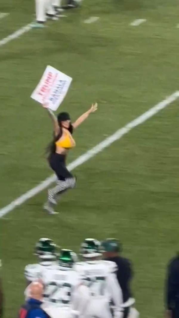 Woman running o<em></em>nto a football field during a game, holding a sign that reads 'Trump, secure border. Kamala open border.'