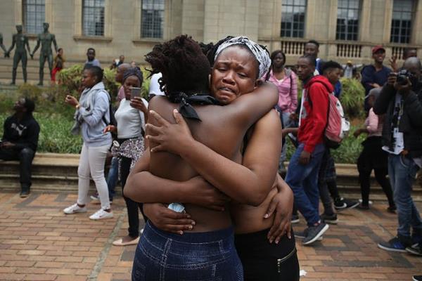 Three women embrace one another, topless and with clothed people passing in the background. One of their faces is facing the camera, she's emotional.