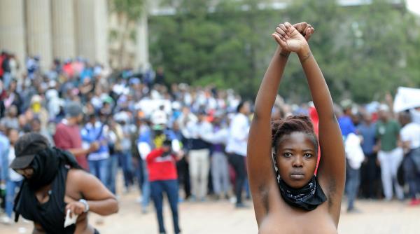 A woman stands, topless with arms raised in the air and crossed as if shackled, a bandana around her neck and a determined look on her face. Behind her, a crowd of people and a cameraman.