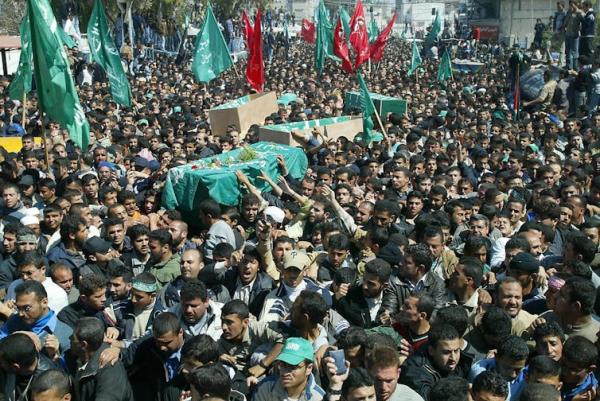 A coffin draped in a green sheet is seen being held by mourners, many waving flags.