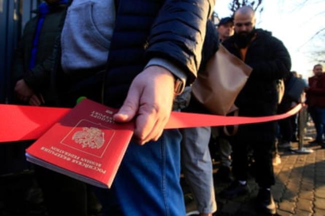 A man holds a Russian passport as he queues with others next to a red cordon