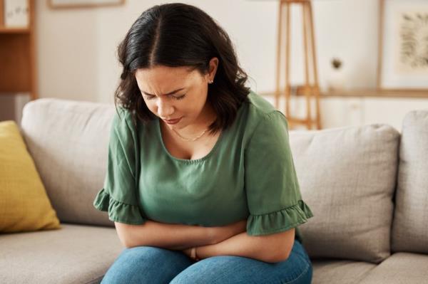Woman in a green blouse sitting on the couch holding her stomach in pain